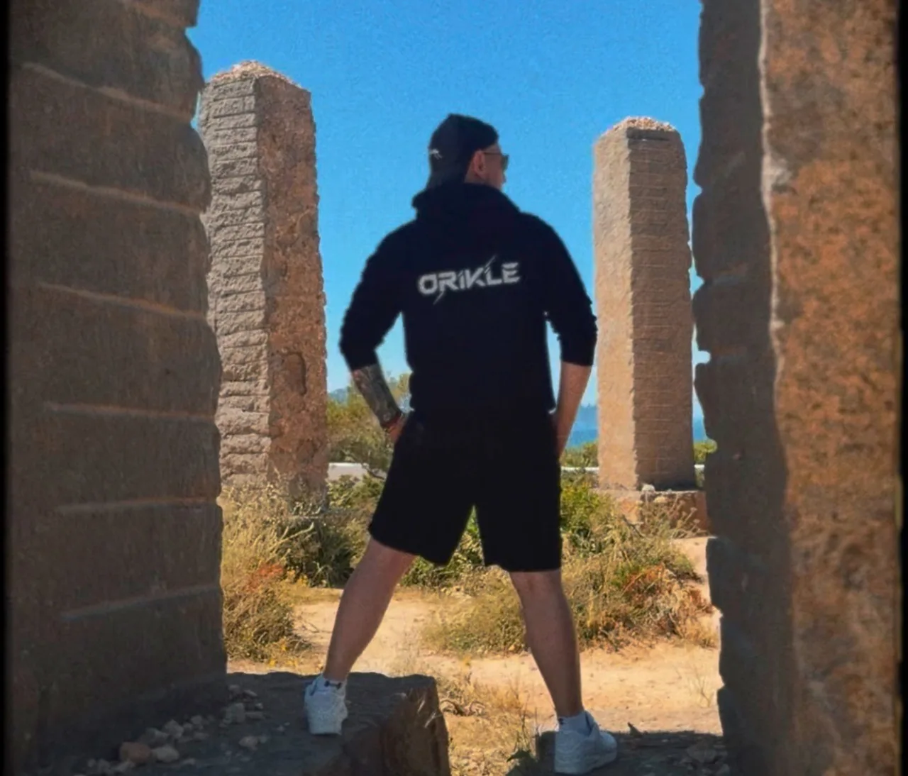 ORIKLE standing between stone pillars in Ibiza ruins, wearing a black ORIKLE hoodie, looking out at the horizon